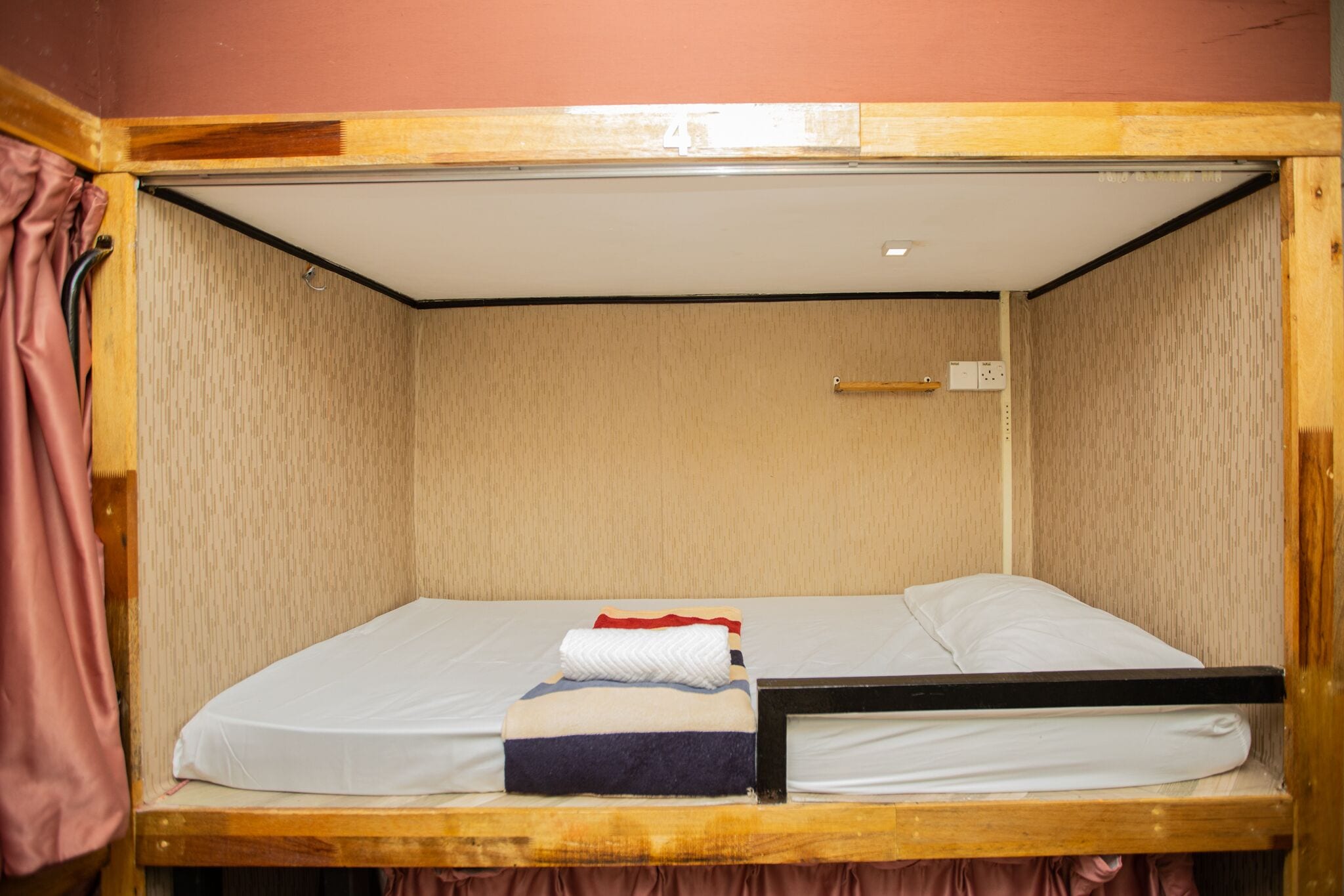 Una cama litera con estructura de madera y colchón blanco. Hay un estante pequeño en la pared y una cortina al lado izquierdo.