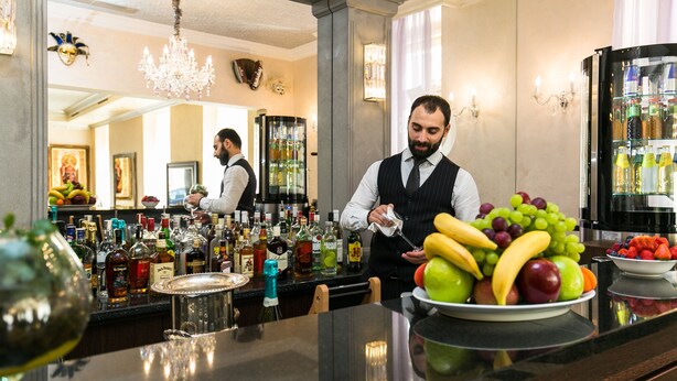 Uma área de bar com diversos tipos de garrafas, uma cesta de frutas e alguém preparando uma bebida.