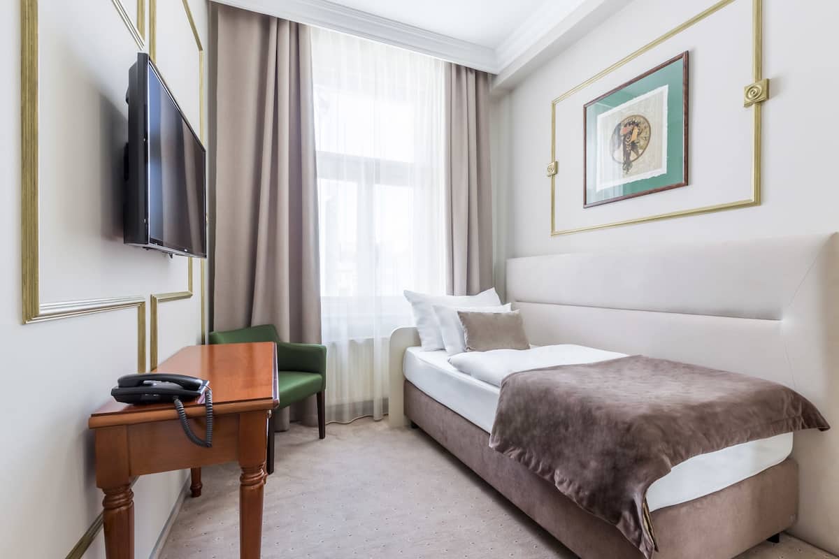 Camera d'albergo con un letto, una scrivania con il telefono, una televisione, una sedia verde e una finestra con tende.