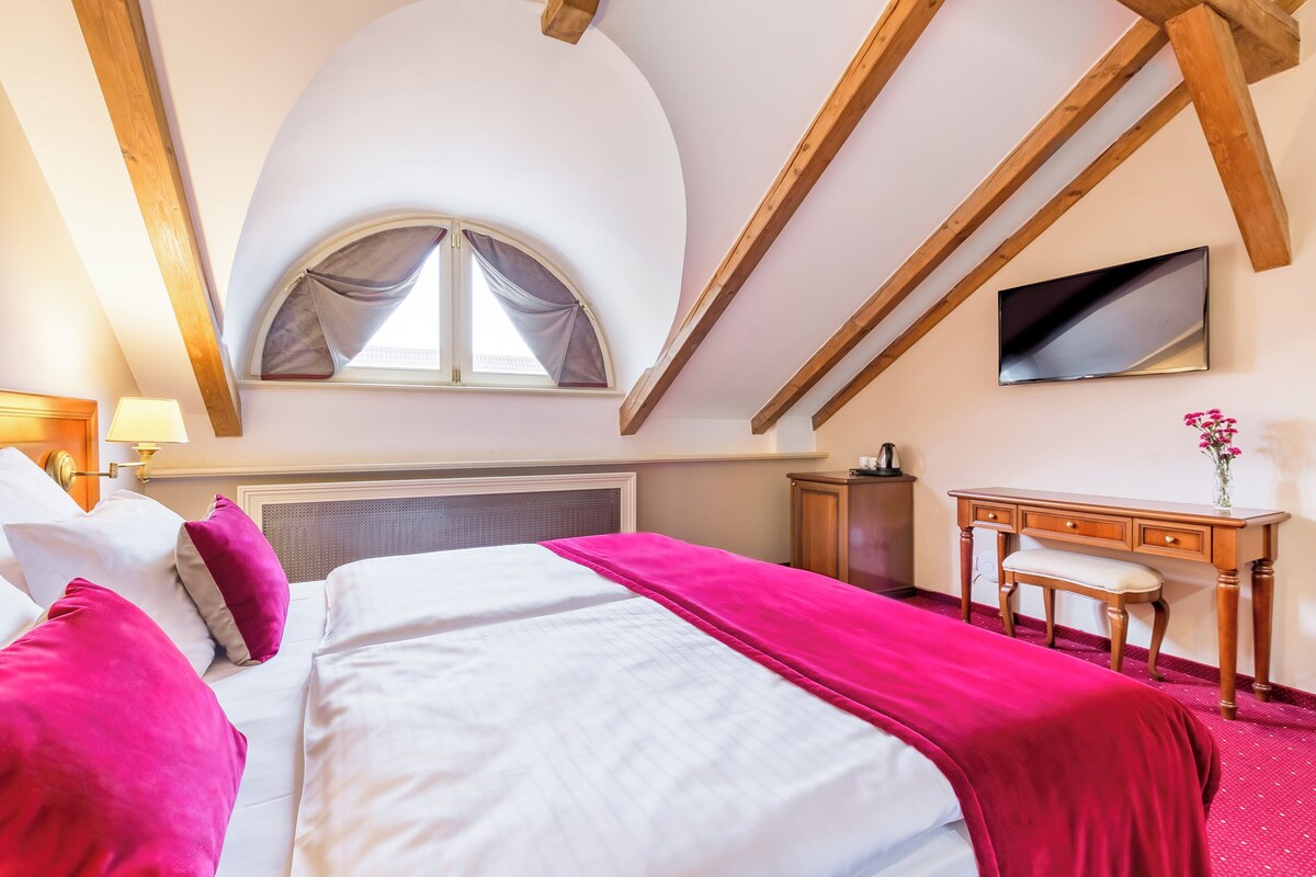 A bedroom with a large bed, a desk, a chair, a TV, and a window with curtains.