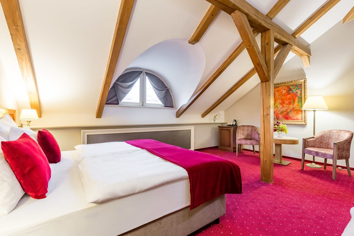 Una camera d'albergo con un letto, una moquette rossa, una finestra con tende, un quadro appeso al muro e un tavolino con una lampada.