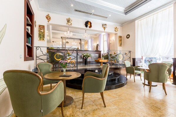 A well-lit, elegant dining area with green upholstered chairs, a marble floor, and a central bar area with a chandelier. - Hotel Caruso Old Town Prague (Prague)