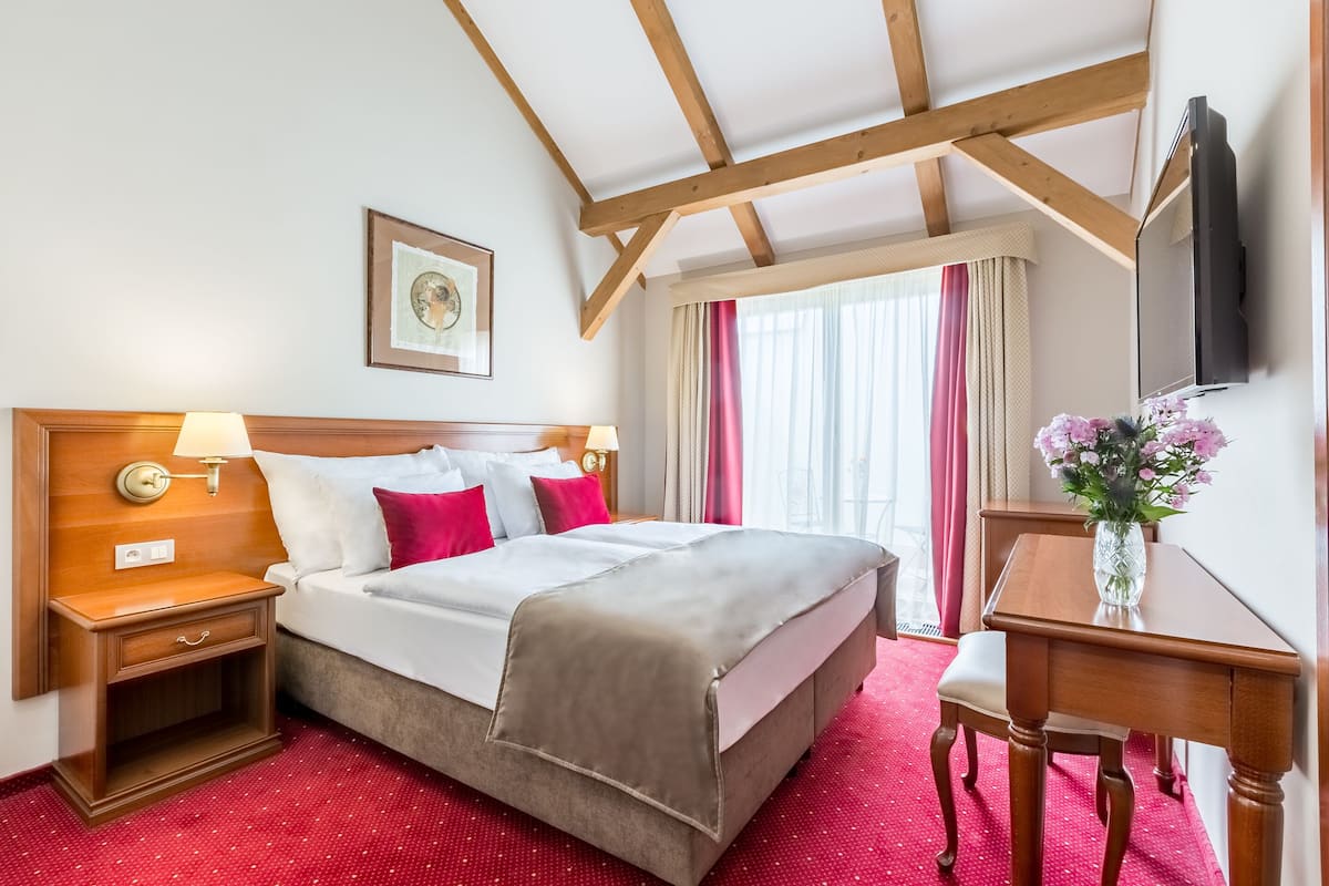 Camera d'albergo con soffitto in legno, un letto con biancheria da letto bianca e cuscini rossi, un comodino con una lampada, una scrivania con un vaso di fiori e una televisione a parete.