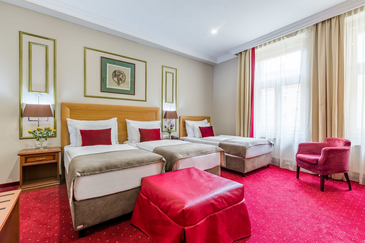 Una camera d'albergo con quattro letti, un poggiapiedi rosso, una poltrona rossa e un'ampia finestra con tende leggere.
