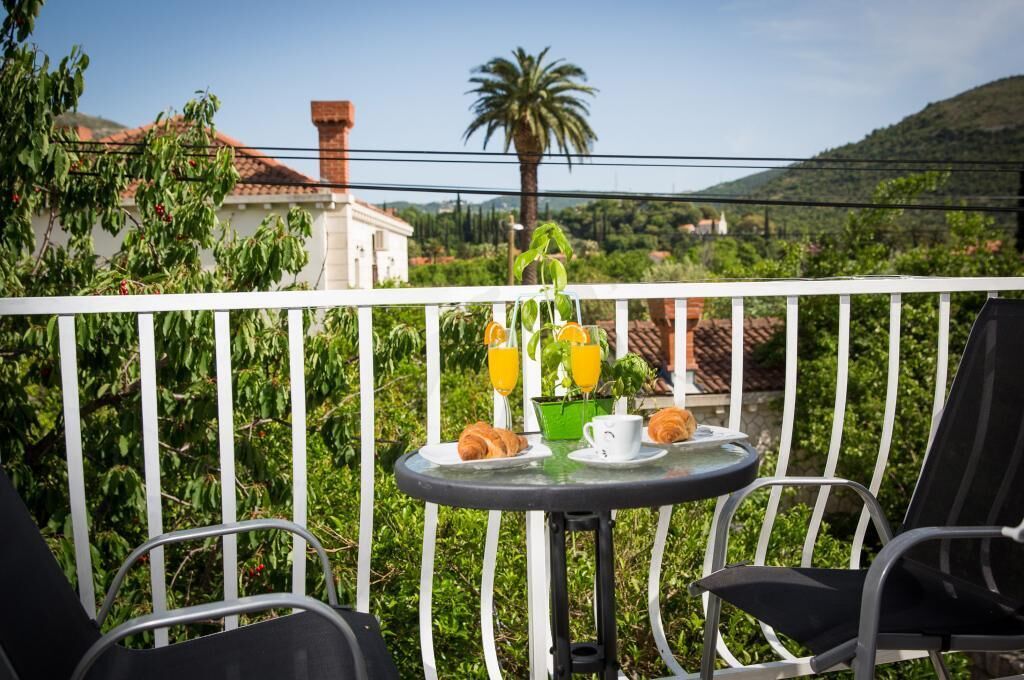 Un balcón con una mesa dispuesta para el desayuno, que incluye cruasanes y jugo de naranja, con vistas a un paisaje pintoresco con colinas y una palmera.