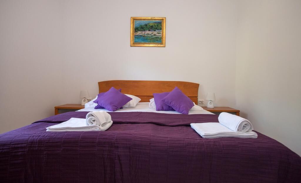 A neatly made bed with purple bedding and pillows, a wooden headboard, and a painting on the wall.