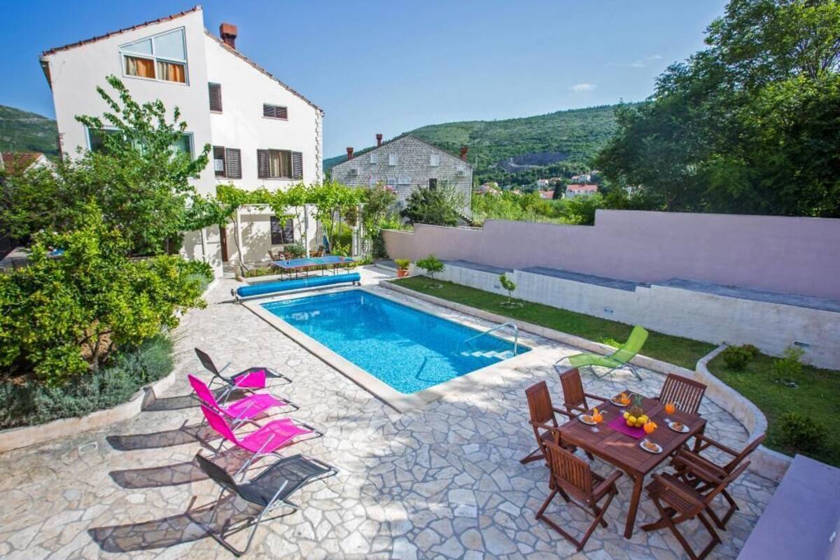 A pool area with lounge chairs and a dining table, surrounded by greenery and a stone patio.