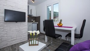Una sala moderna con televisor de pantalla plana, comedor con mesa y sillas, y cocina al fondo.