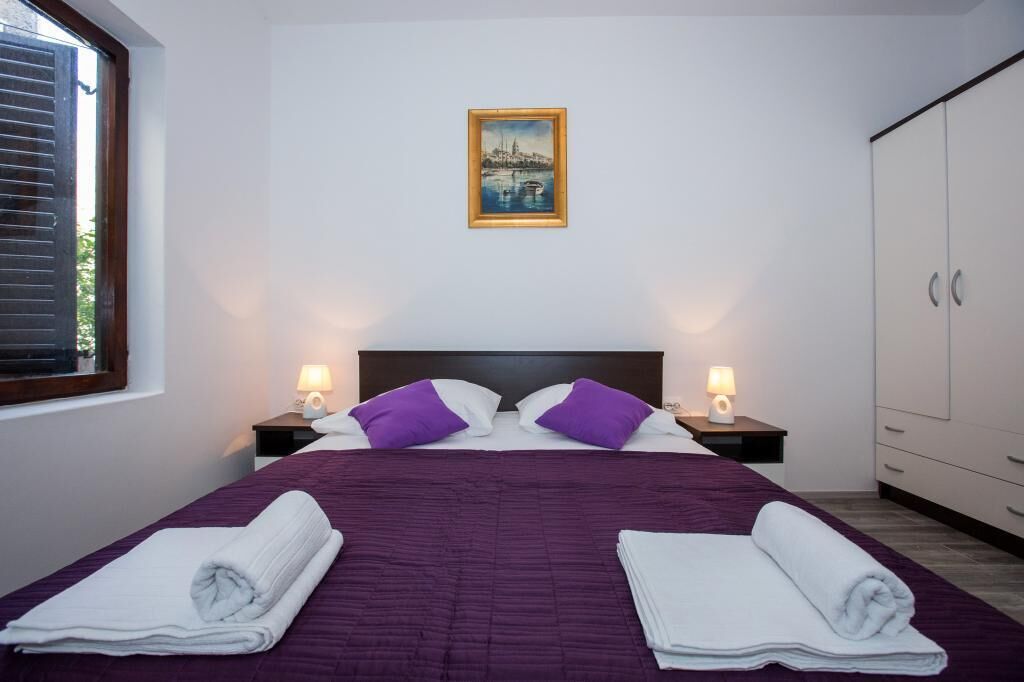 A neatly made bed with purple bedding and white towels, a painting above the bed, and a wardrobe to the right.