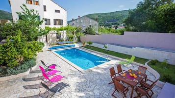 A pool area with lounge chairs and a dining table, surrounded by greenery and a stone patio.