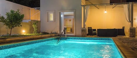 A swimming pool with a tiled edge, a covered seating area, and a building with a door leading to an interior space.
