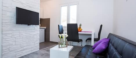 Ein modernes Wohnzimmer mit einer schwarzen Ledersofa, einem weißen Couchtisch und einem Flachbildfernseher, der an einer weißen Ziegelwand angebracht ist. Es gibt einen Essbereich mit Tisch und Stühlen, und im Hintergrund befindet sich eine Küche.