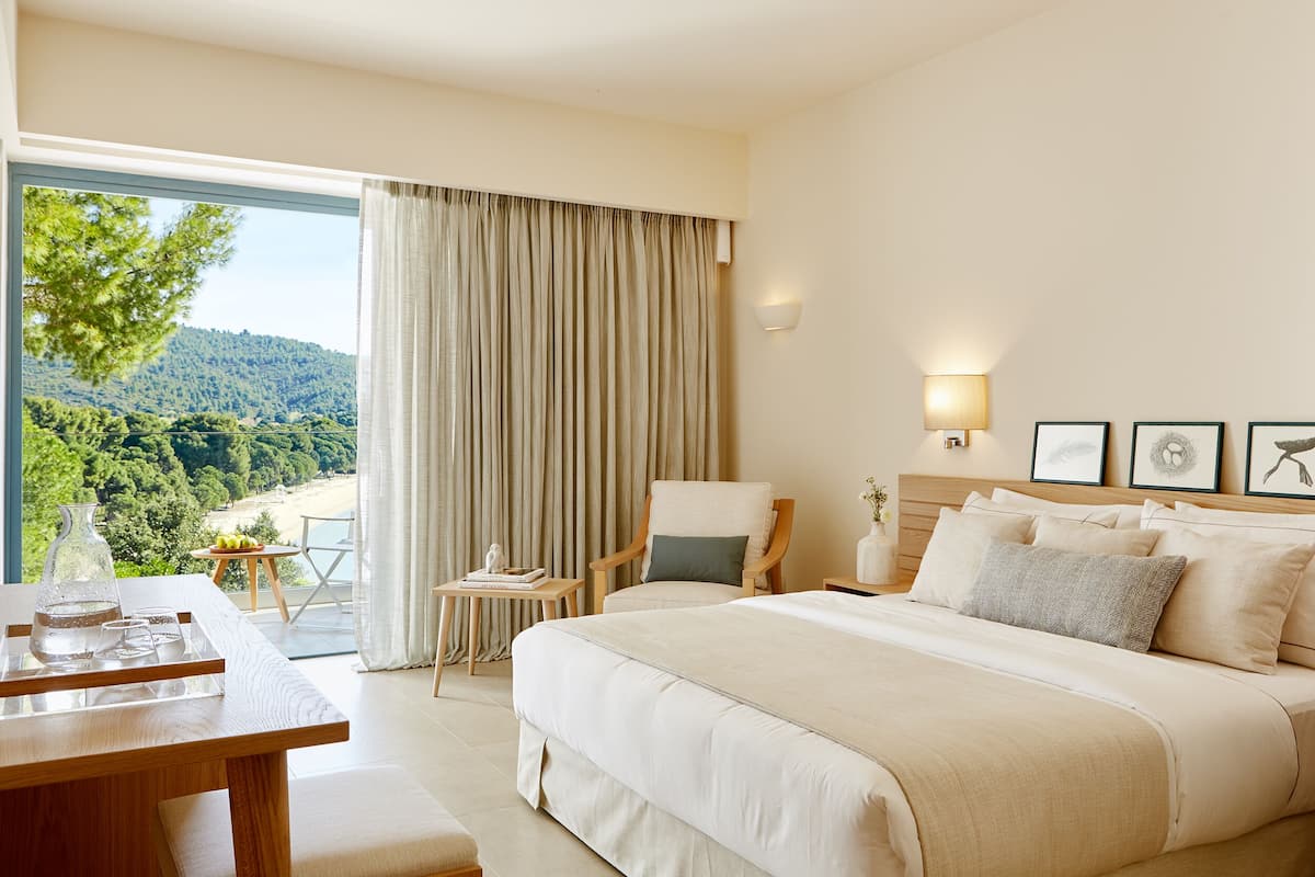 A bedroom with a large bed, a chair, a table with a vase, and a view of a forested area.