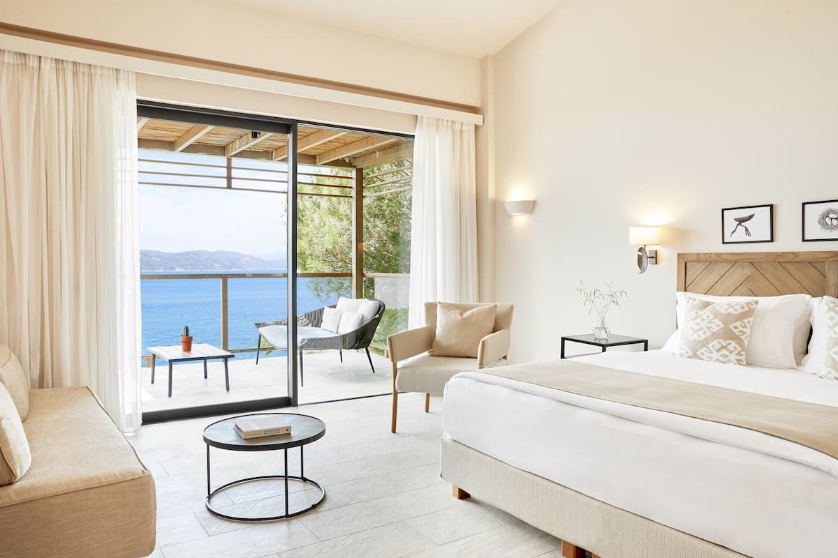 A bedroom with a large bed, a sofa, a round coffee table, and a view of the sea through a sliding glass door.