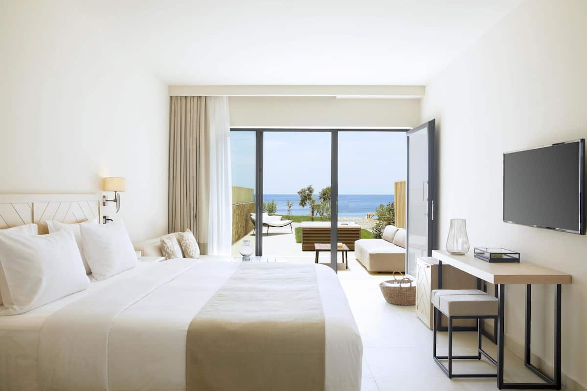 A hotel room with a large bed, a desk, a TV, and a balcony with a view of the ocean.