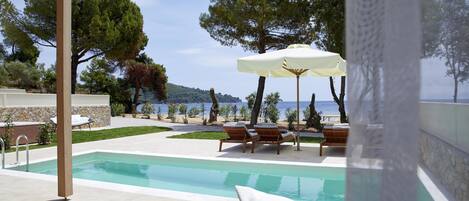 A pool area with lounge chairs and an umbrella, surrounded by trees and a view of the sea.