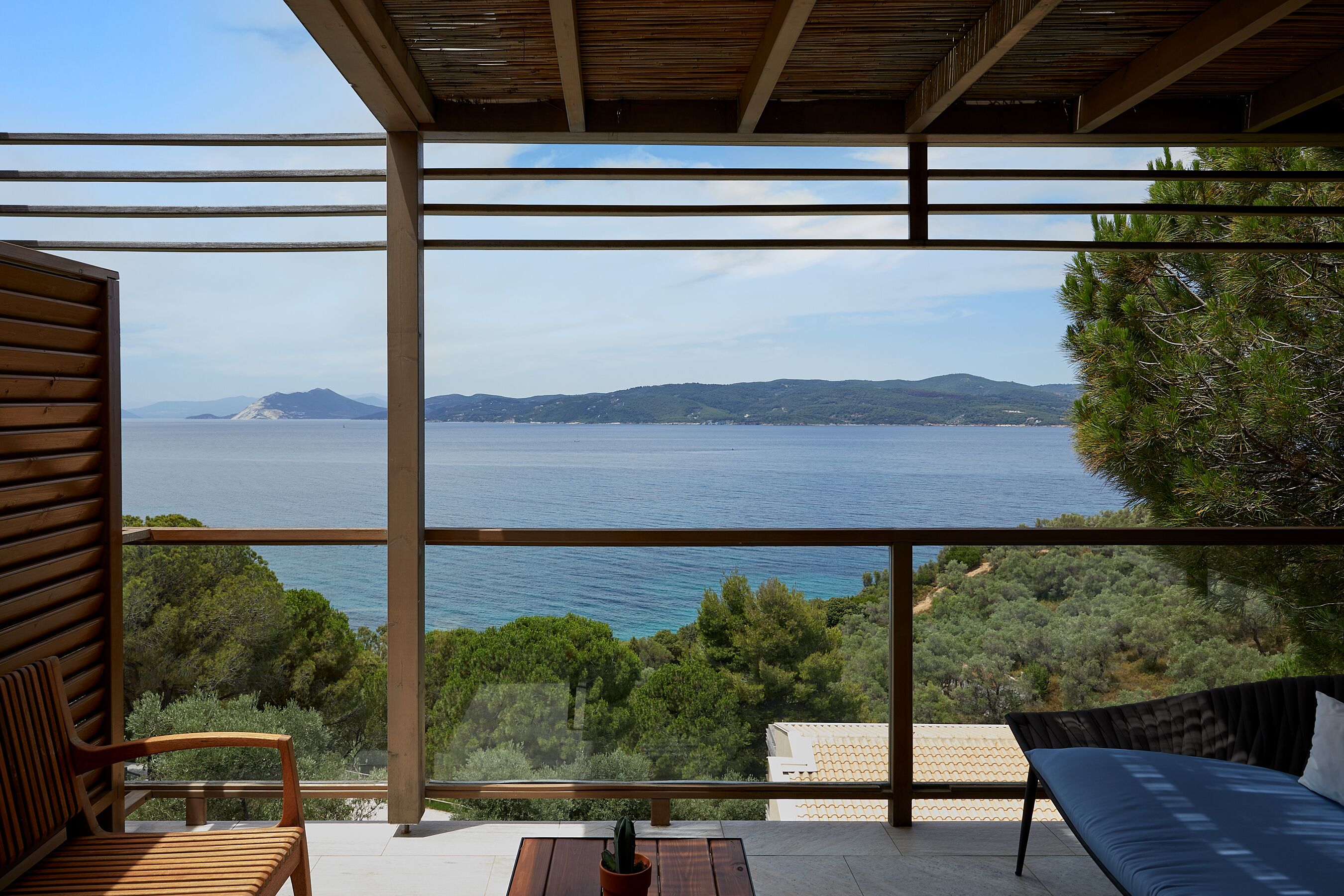 Un balcón con vista al mar y colinas, muebles de madera y una planta en maceta.