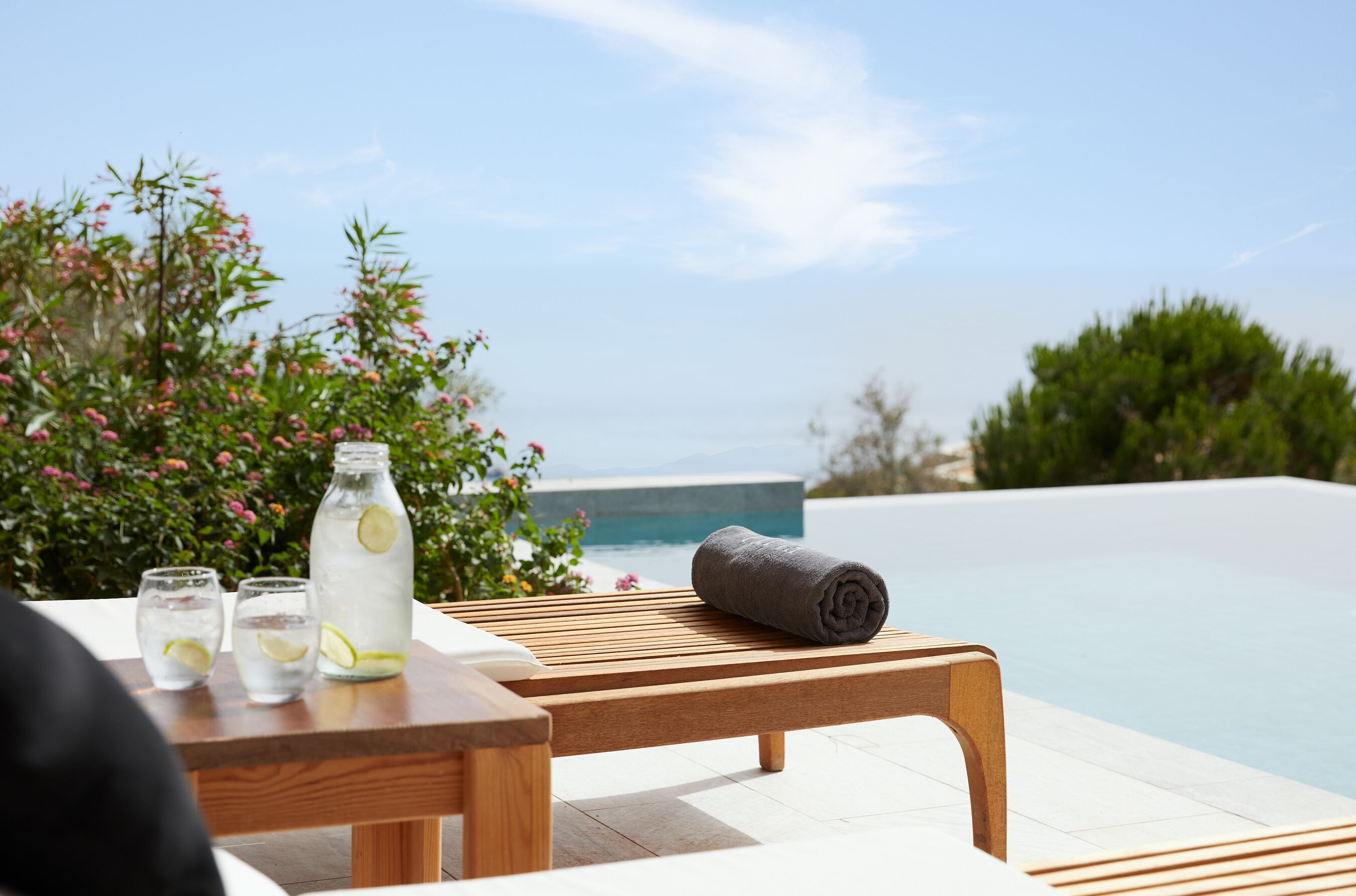 Ein Holztisch mit einer Wasserflasche und Gläsern, einem aufgerollten Handtuch und Blick auf einen Swimmingpool und Grünflächen.