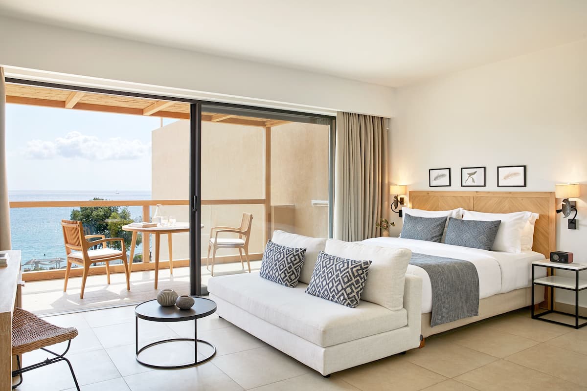 A hotel room with a bed, a sofa, a small table, and a chair. There is a balcony with a view of the sea.