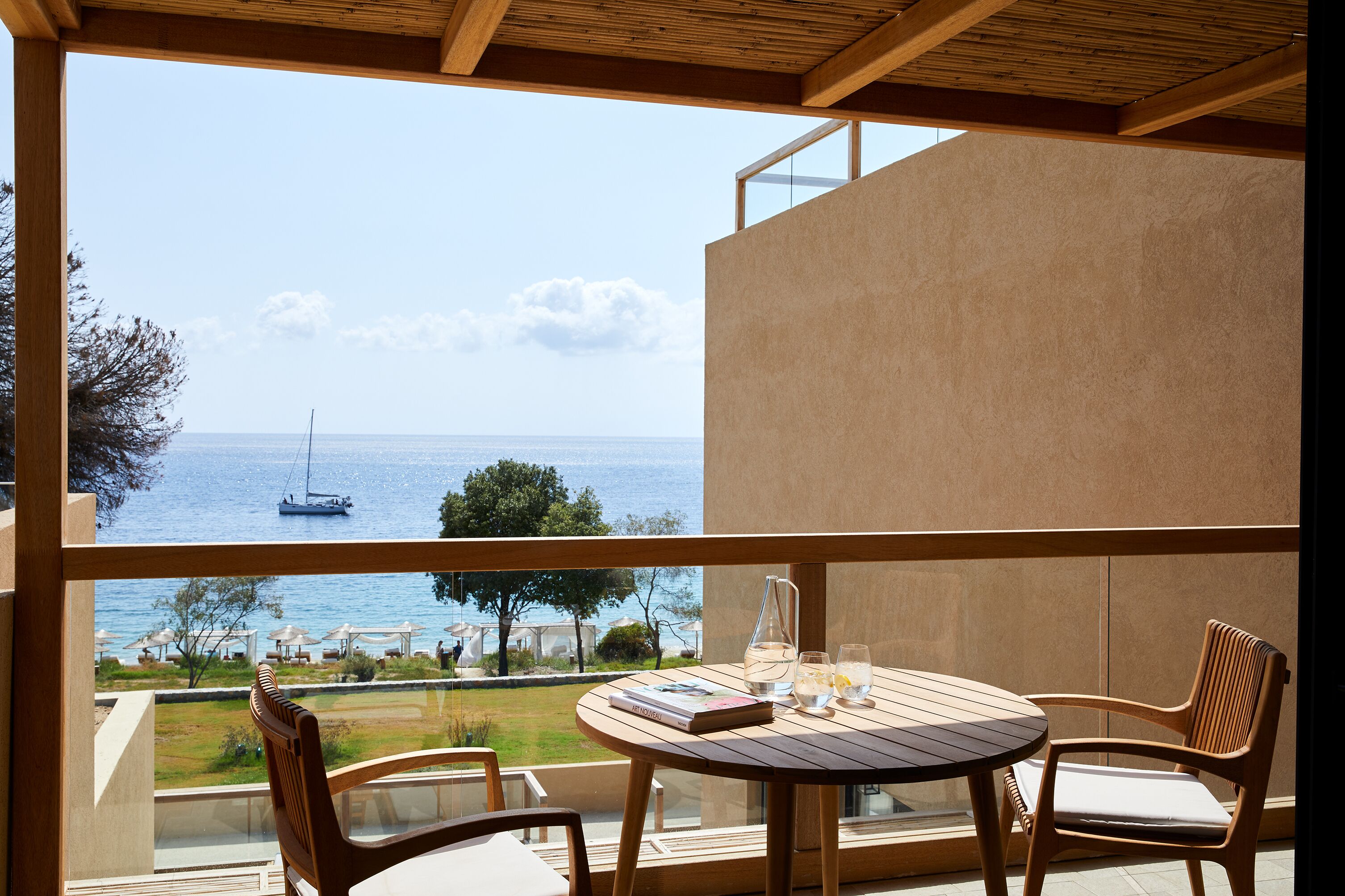 A balcony with a wooden table and chairs, overlooking a beach with umbrellas and a sailboat.