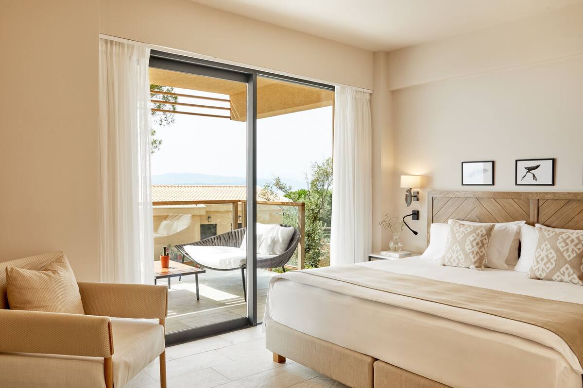 A hotel room with a bed, a chair, a table, and a view of the outdoors.
