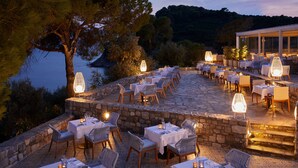 An outdoor dining area with stone flooring, white tablecloths, and wicker chairs, overlooking a body of water and hills. - ELIVI Skiathos (Skiathos)