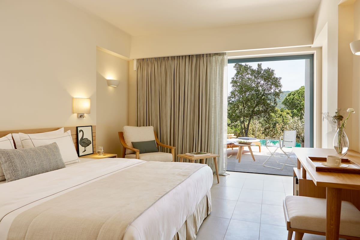 A hotel room with a large bed, a chair, a small table, and a view of the outdoors.