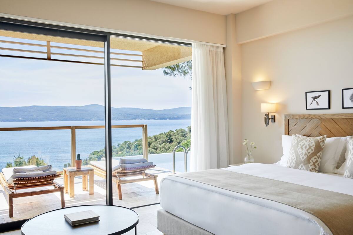 A hotel room with a large bed, a view of the sea, a balcony with seating, and a wooden headboard.