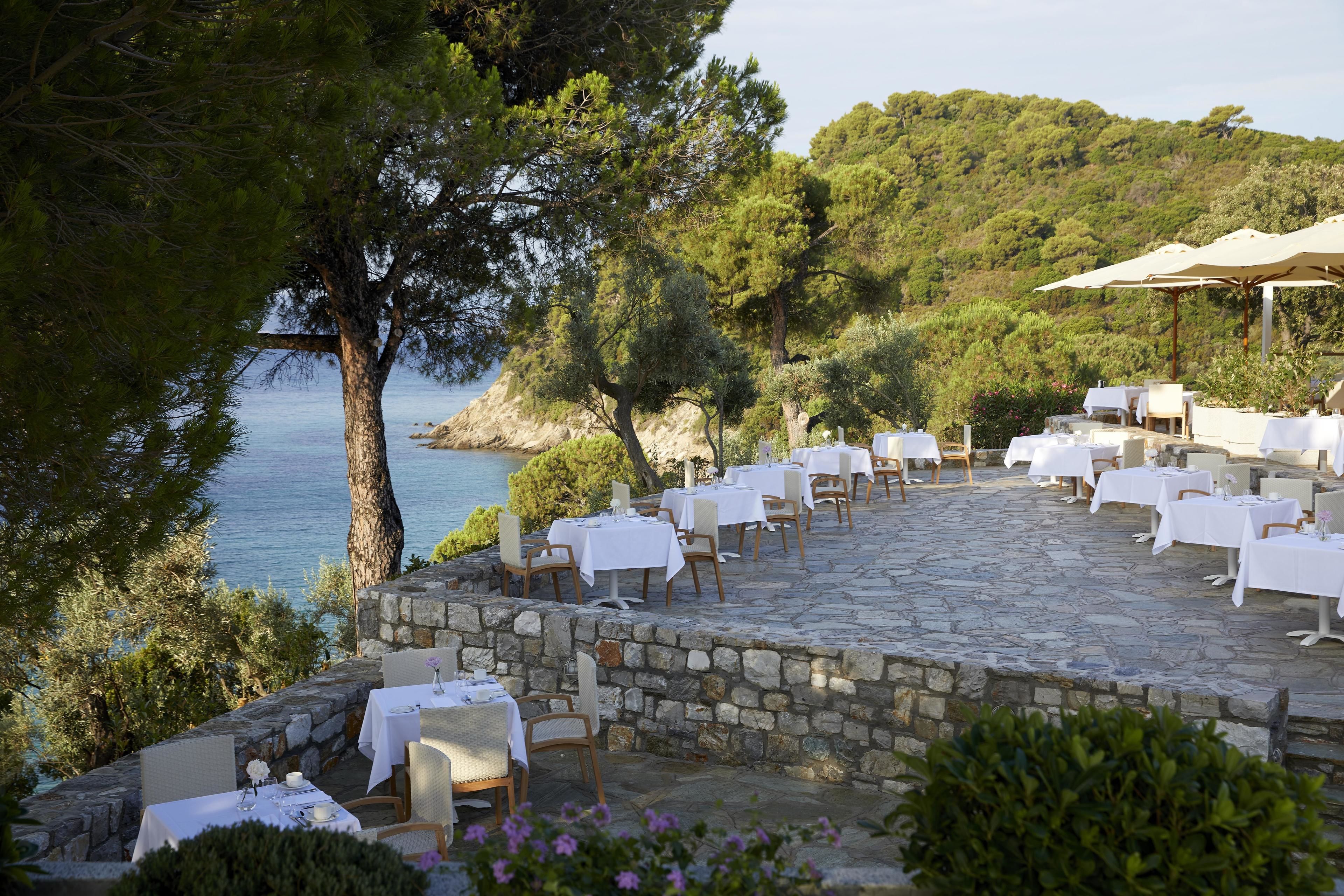 An outdoor dining area with stone flooring, white tablecloths, and chairs, overlooking a body of water and surrounded by trees.