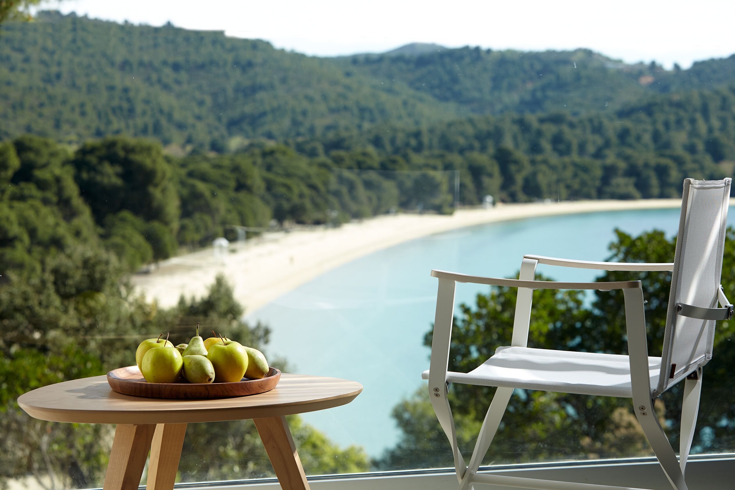 A table with a bowl of apples and pears, a white chair, and a view of a beach and forest.