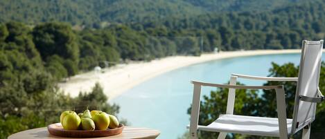 A table with a bowl of apples and pears, a white chair, and a view of a beach and forest.