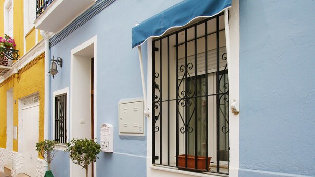 Una calle estrecha con un edificio azul que tiene una ventana con barrotes, una puerta blanca y un pequeño balcón.