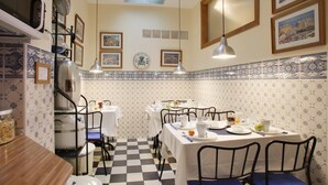 Un comedor con suelo a cuadros, mesas y sillas blancas. Hay cuadros en las paredes y un reloj.