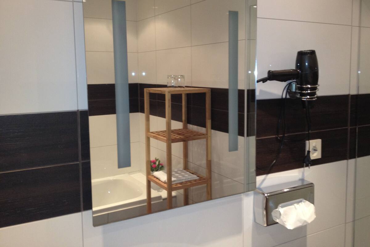 A bathroom with a glass-enclosed shower, a hairdryer on the wall, and a small wooden shelf with a towel and flowers.