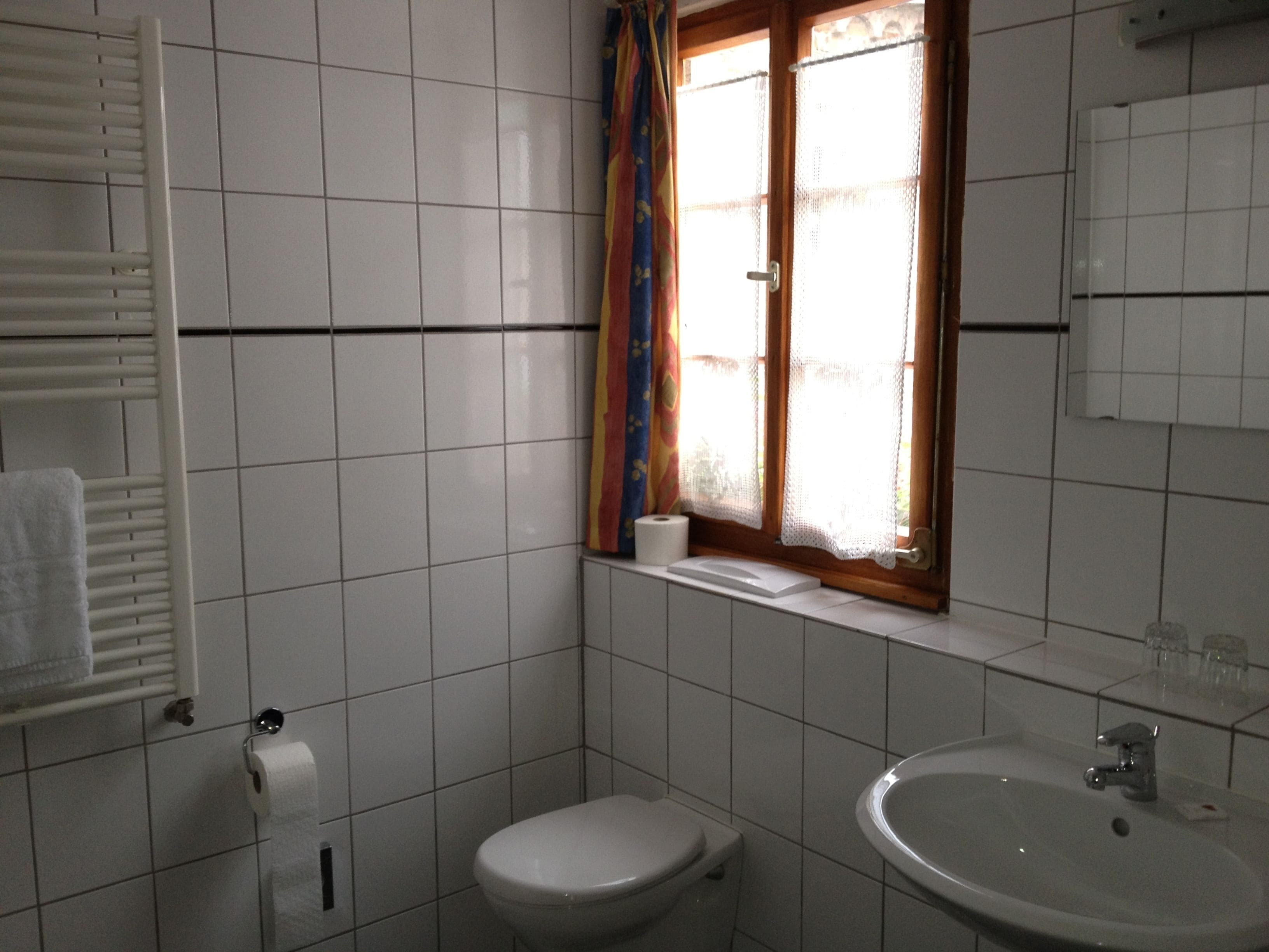 Une salle de bain avec des carreaux blancs, des toilettes, un lavabo, une fenêtre avec des rideaux et un porte-serviettes.