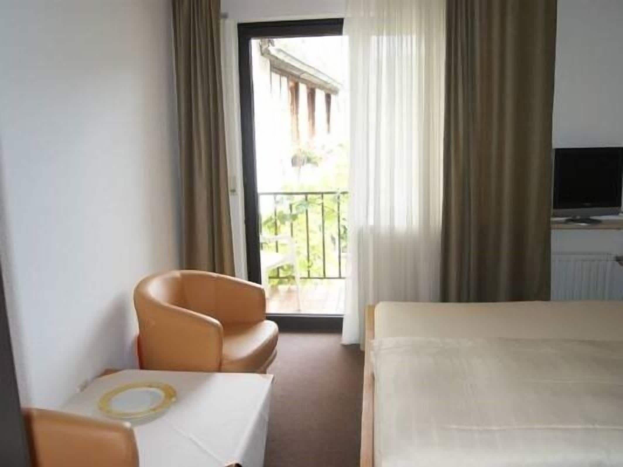 Ein Hotelzimmer mit Bett, Sessel, kleinem Tisch, Fernseher und Balkon mit Vorhängen.