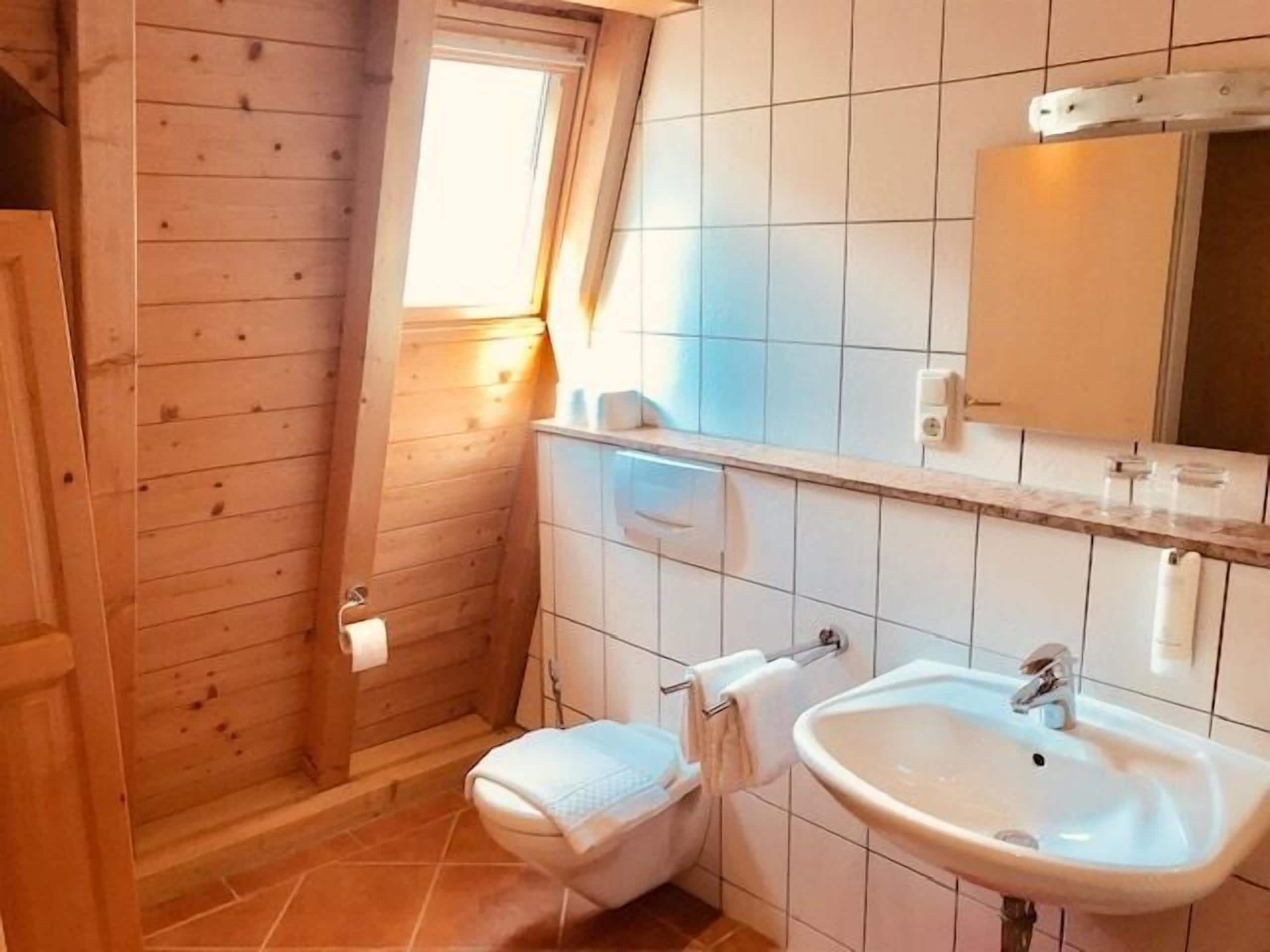 Une petite salle de bain avec une porte en bois, une fenêtre, un lavabo blanc et un miroir.