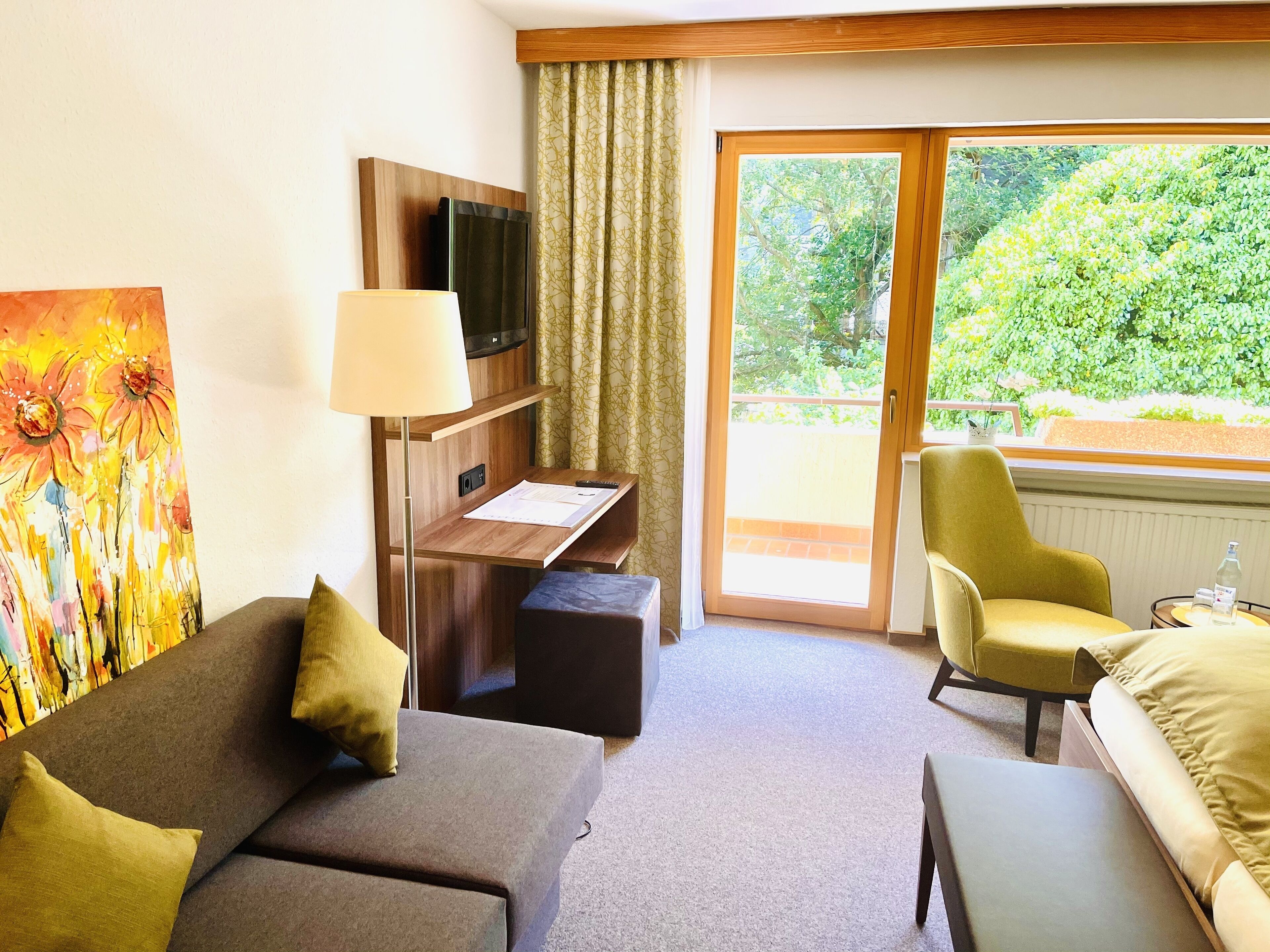 Ein Hotelzimmer mit einer Couch, einem Schreibtisch mit Lampe, einem Fernseher und Blick auf Grünflächen durch das Fenster.
