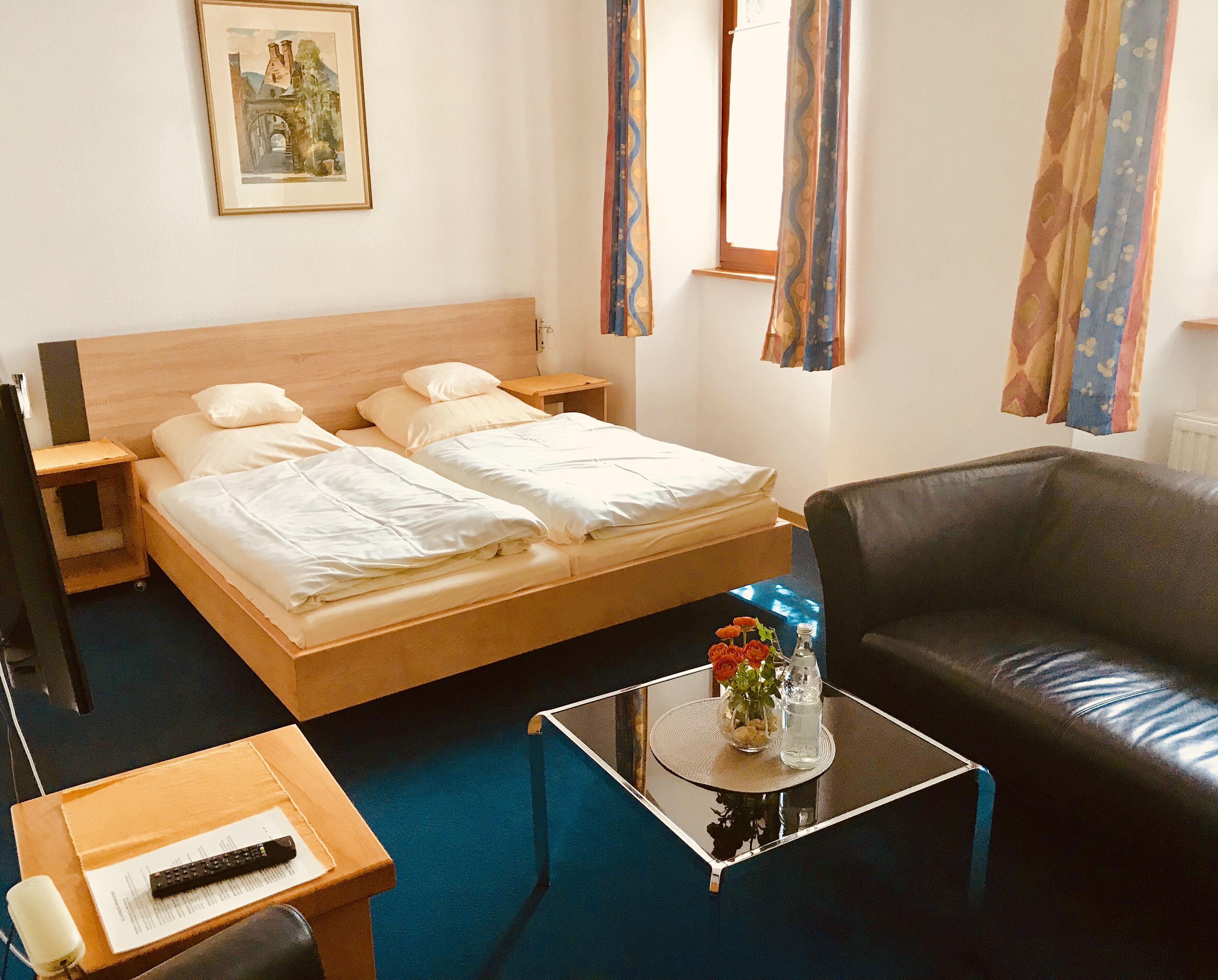 Une chambre d’hôtel avec un lit double, une petite table avec une télécommande, une table basse ornée de fleurs et un canapé en cuir.