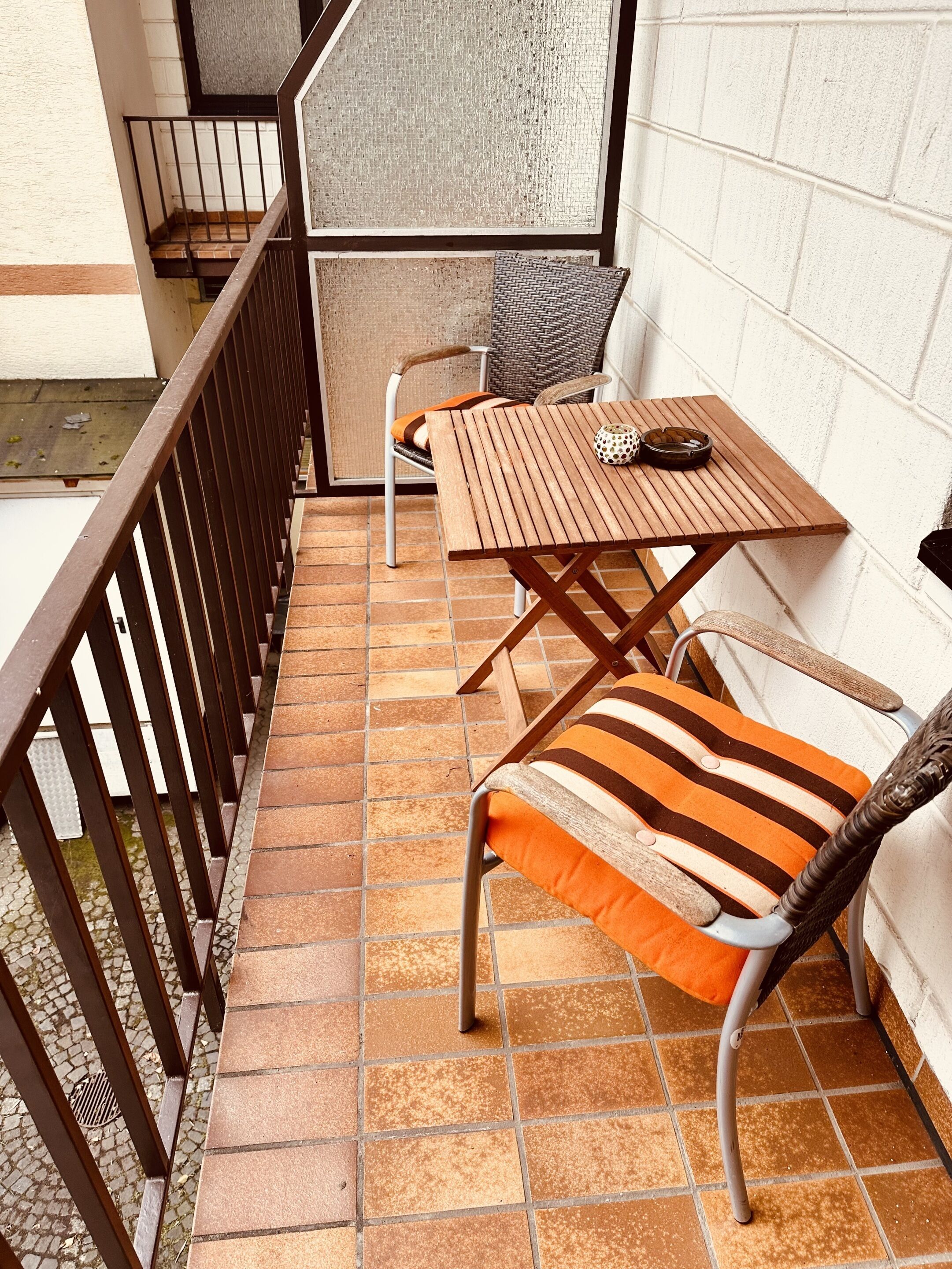 A small outdoor patio with a wooden table and two chairs, a tiled floor, and a metal railing.