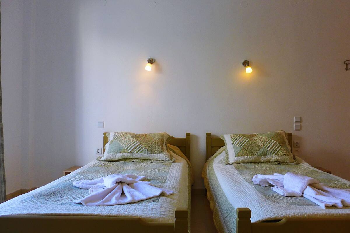 Dos camas individuales con cabeceros, cada una con colcha acolchada y almohadas. Hay lámparas fijadas a la pared sobre las camas.