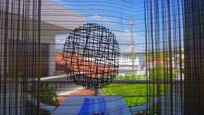 Vista a través de una ventana con un diseño de cuadrícula, posiblemente una malla o persianas, con un fondo desenfocado que incluye un edificio y follaje.