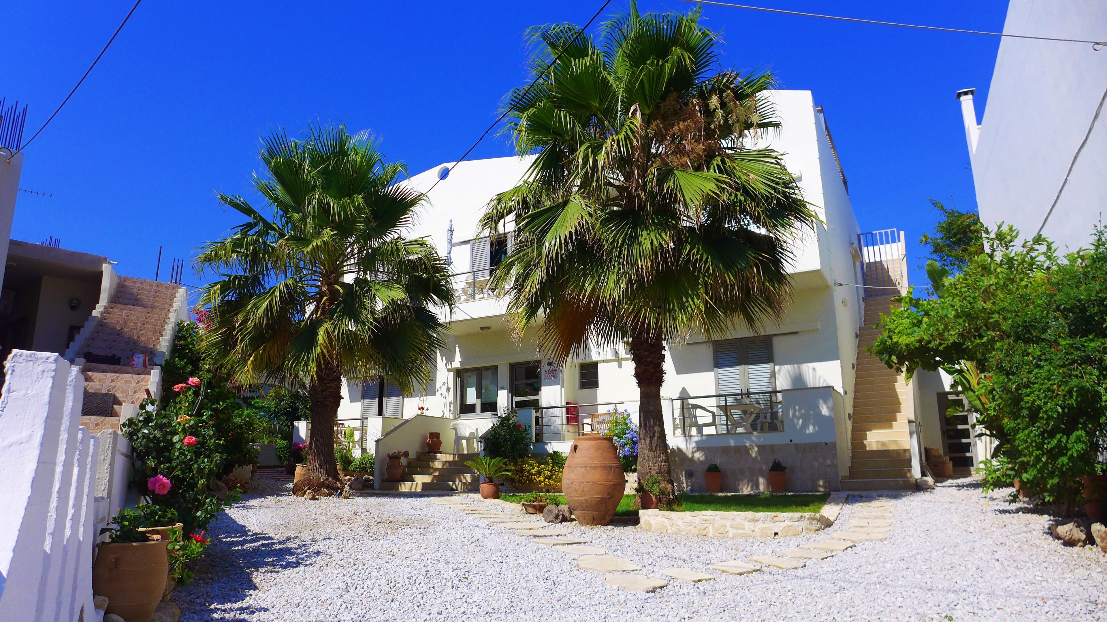 A two-story building with a white facade, a balcony, and a gravel courtyard with palm trees and potted plants.