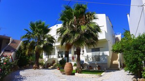 A white building with a balcony, surrounded by palm trees and a gravel pathway. - Lion-Suites (Apokoronas)