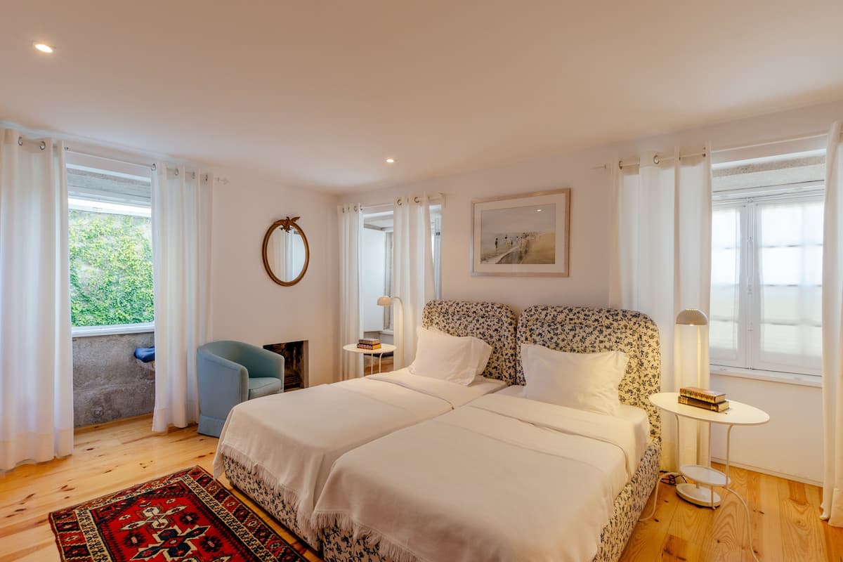 A bedroom with two beds, a small table, a chair, a mirror, and a window with curtains.