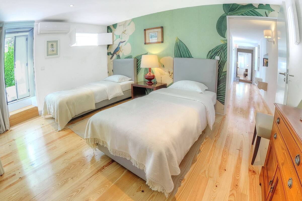 A hotel room with two beds, a wooden floor, a green leaf mural, a nightstand with a lamp, and a wooden dresser.