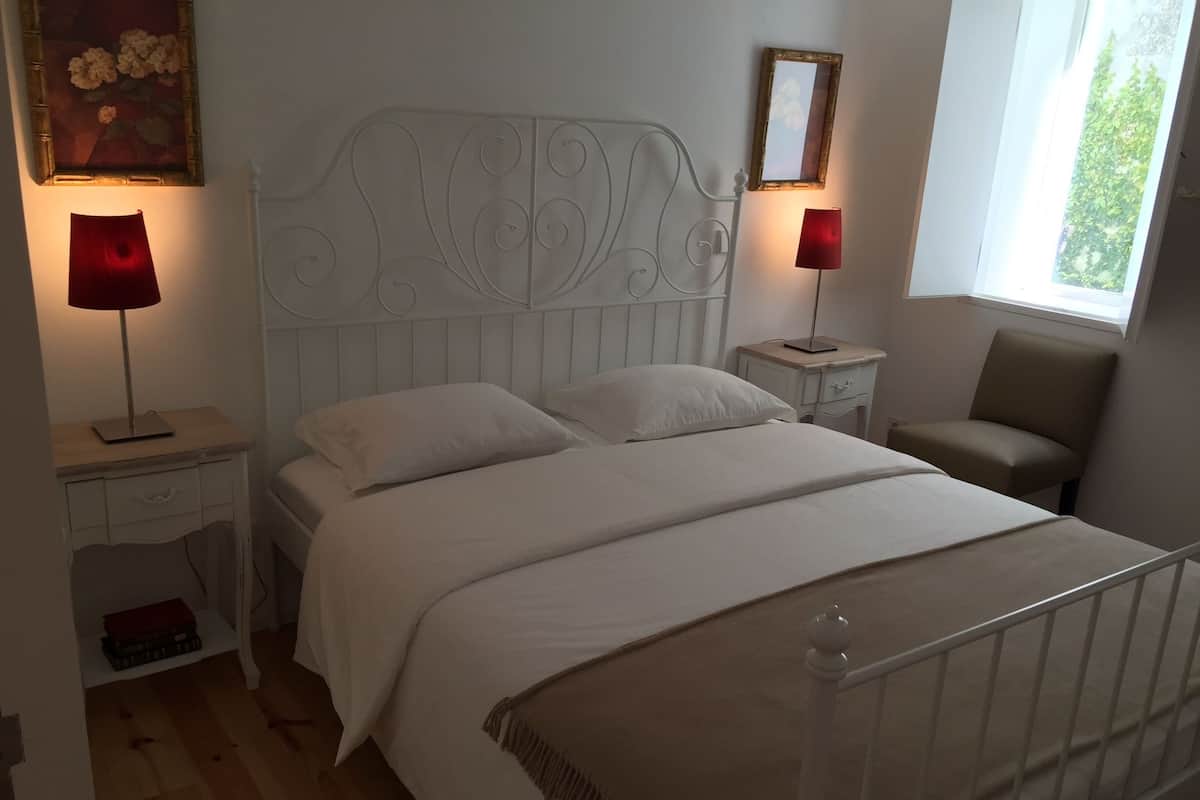 A bedroom with a white bed, two bedside tables with lamps, a chair, and a window.