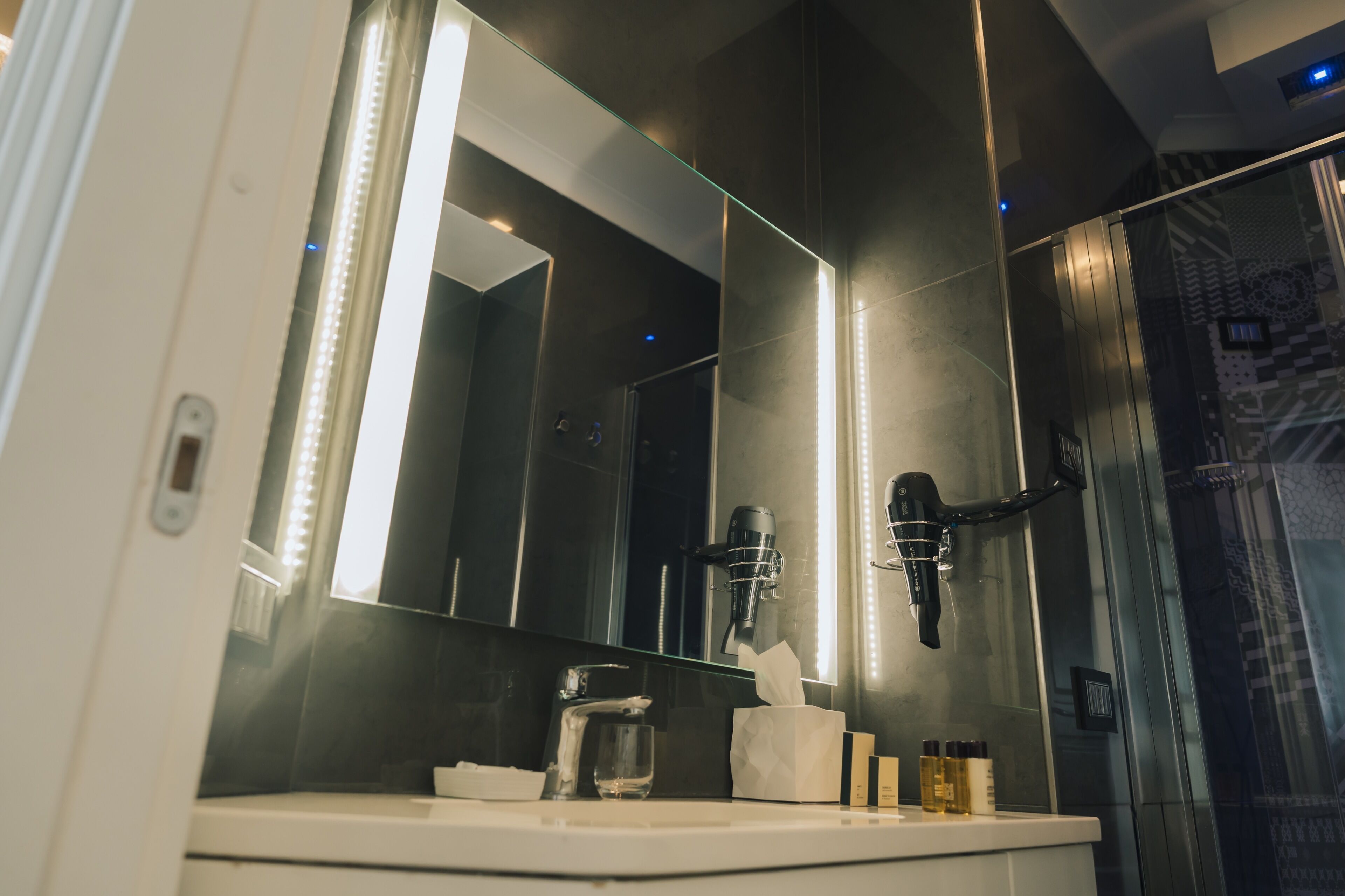 A modern bathroom with a large mirror, a hairdryer, a glass of water, and toiletries on the counter.
