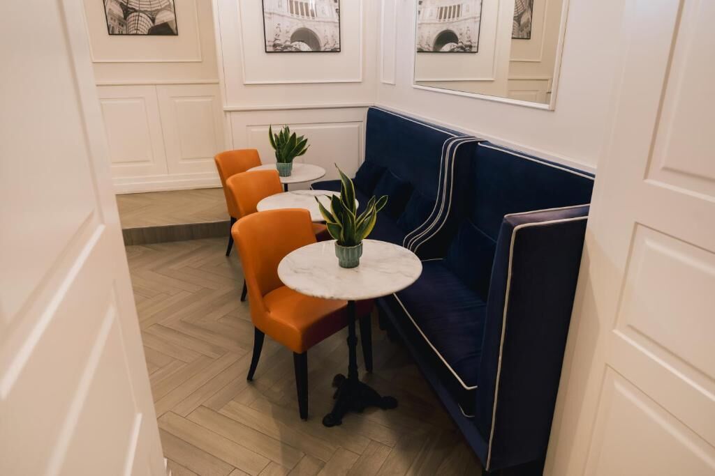 A corner seating area with a blue cushioned bench, a round table with a potted plant, and two orange chairs.