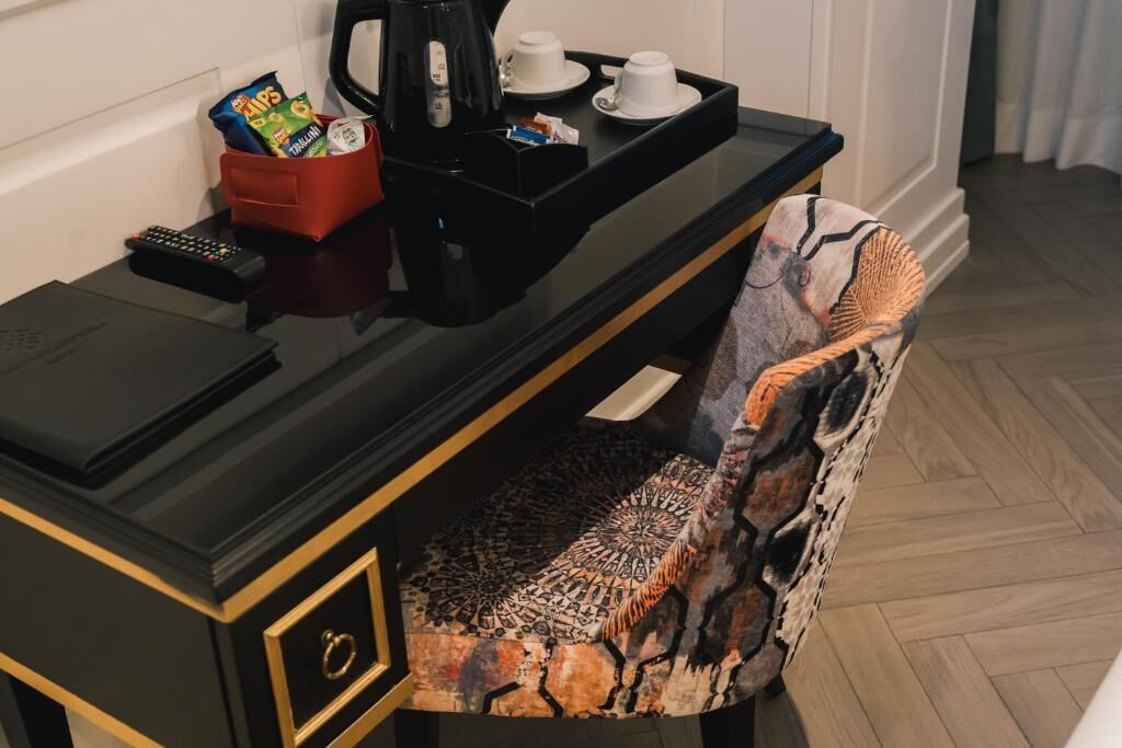 A hotel room with a black desk, a red snack container, a coffee maker, and a patterned chair.